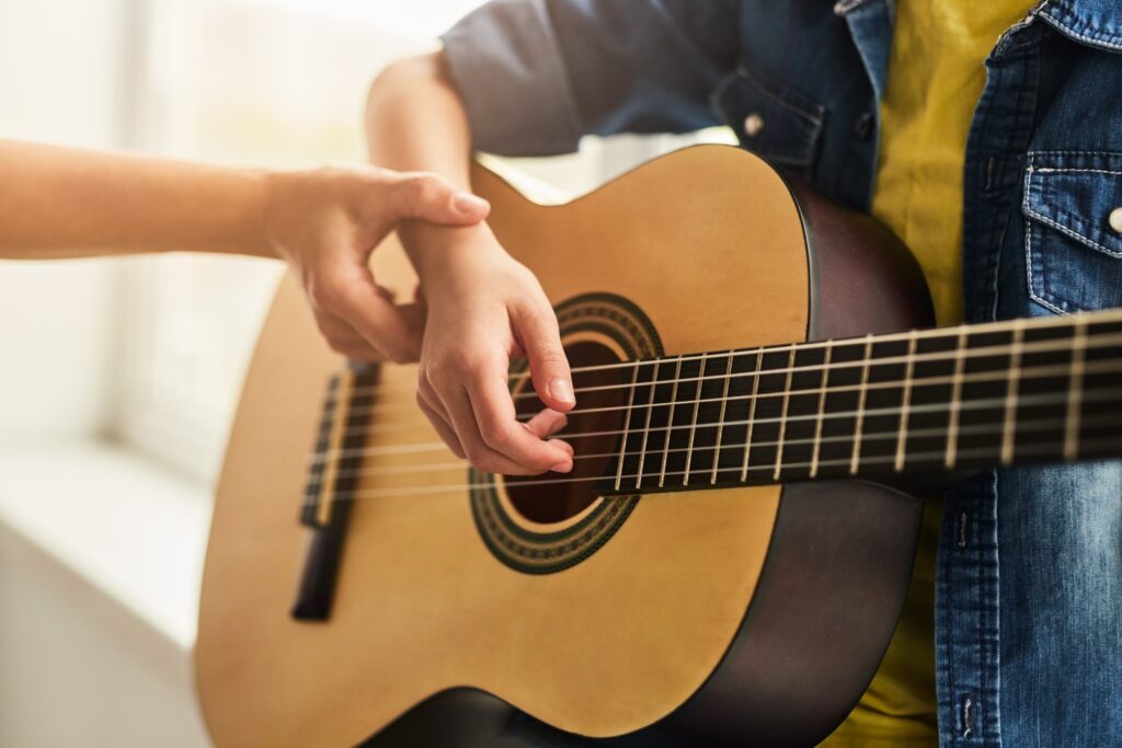 Unrecognizable tutor adjusting hand of teenager on strings of acoustic guitar during lesson in music school in daytime